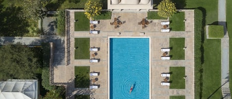 An aerial view of a rectangular swimming pool surrounded by lounge chairs and tables, with a covered seating area and a small boat in the pool.