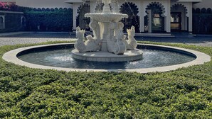 Ein Brunnen mit einer zentralen Statue, umgeben von einem kreisförmigen Becken und üppigem Grün. Im Hintergrund ist ein Gebäude mit einer Kuppel und Bogenfenstern zu sehen.