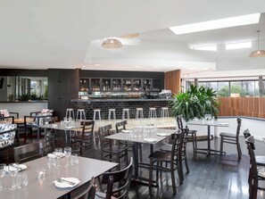 A modern restaurant with high tables and bar stools, a bar area with shelves of bottles, and large windows offering a view. - Mantra Sun City (Surfers Paradise)