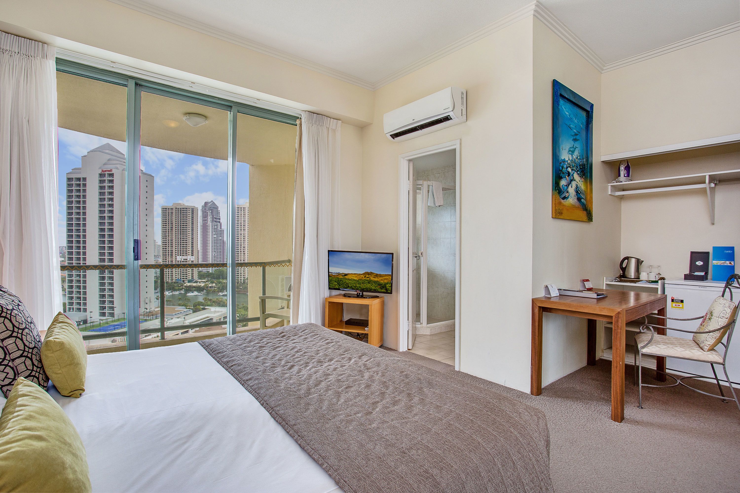 Habitación de hotel con una cama grande, un escritorio con silla, televisión y vistas a edificios de la ciudad a través del ventanal.