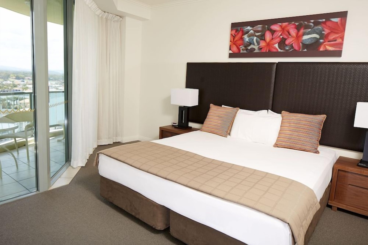 A hotel room with a large bed, two bedside tables, a view of the city through a balcony, and a decorative wall piece.