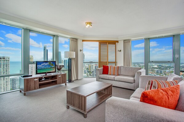 Relax in this stylish living room with a large flat-screen TV, sofa, and floor lamp, offering a panoramic view of the city and ocean.