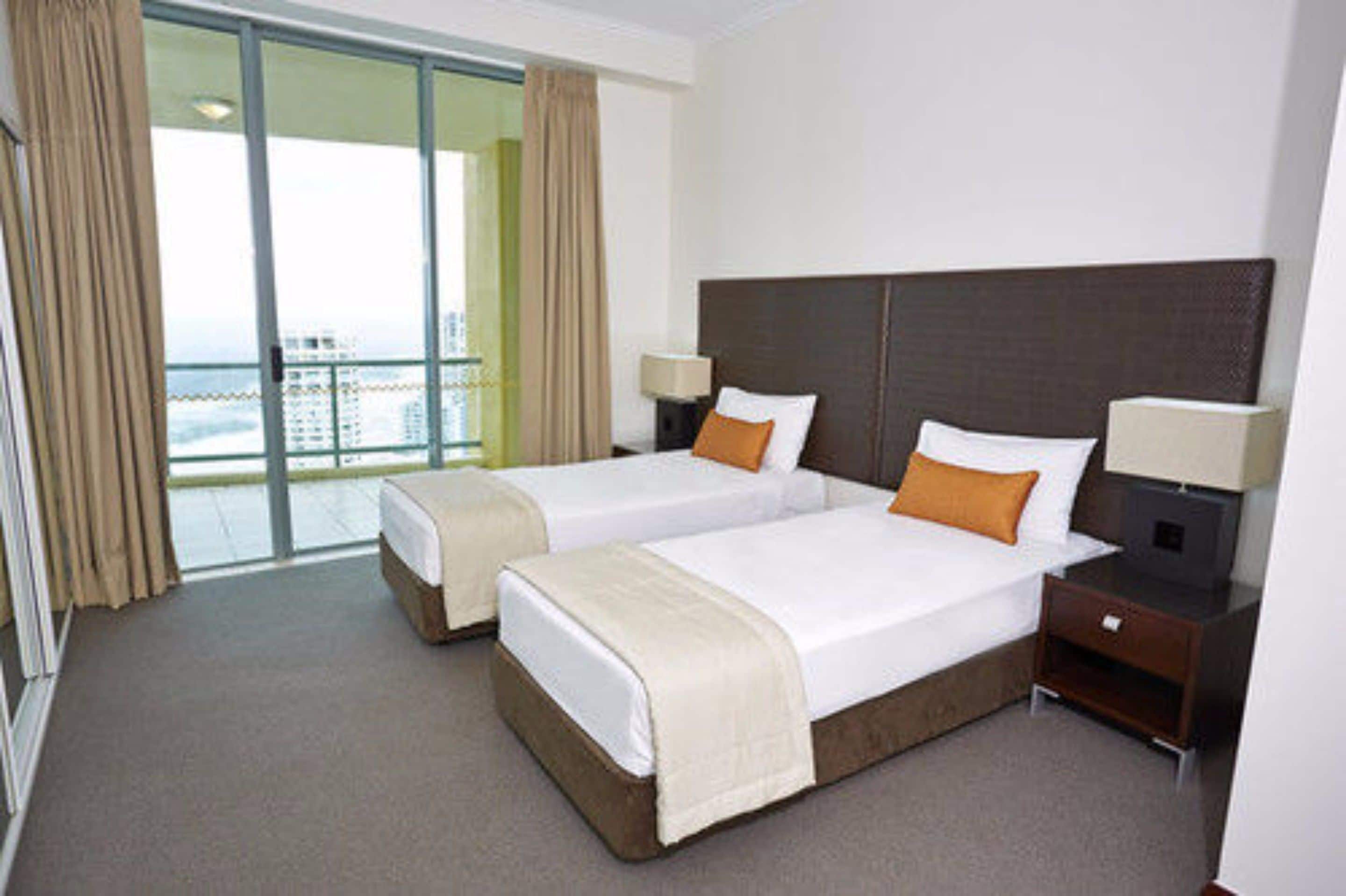 A hotel room with two beds, a balcony with curtains, and a view of the city.