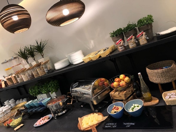 A buffet setup with various food items, including fruits, cheeses, and condiments, arranged on a table with overhead lighting.