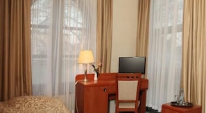 A hotel room with a bed, a desk with a television, a lamp, and a vase with flowers.