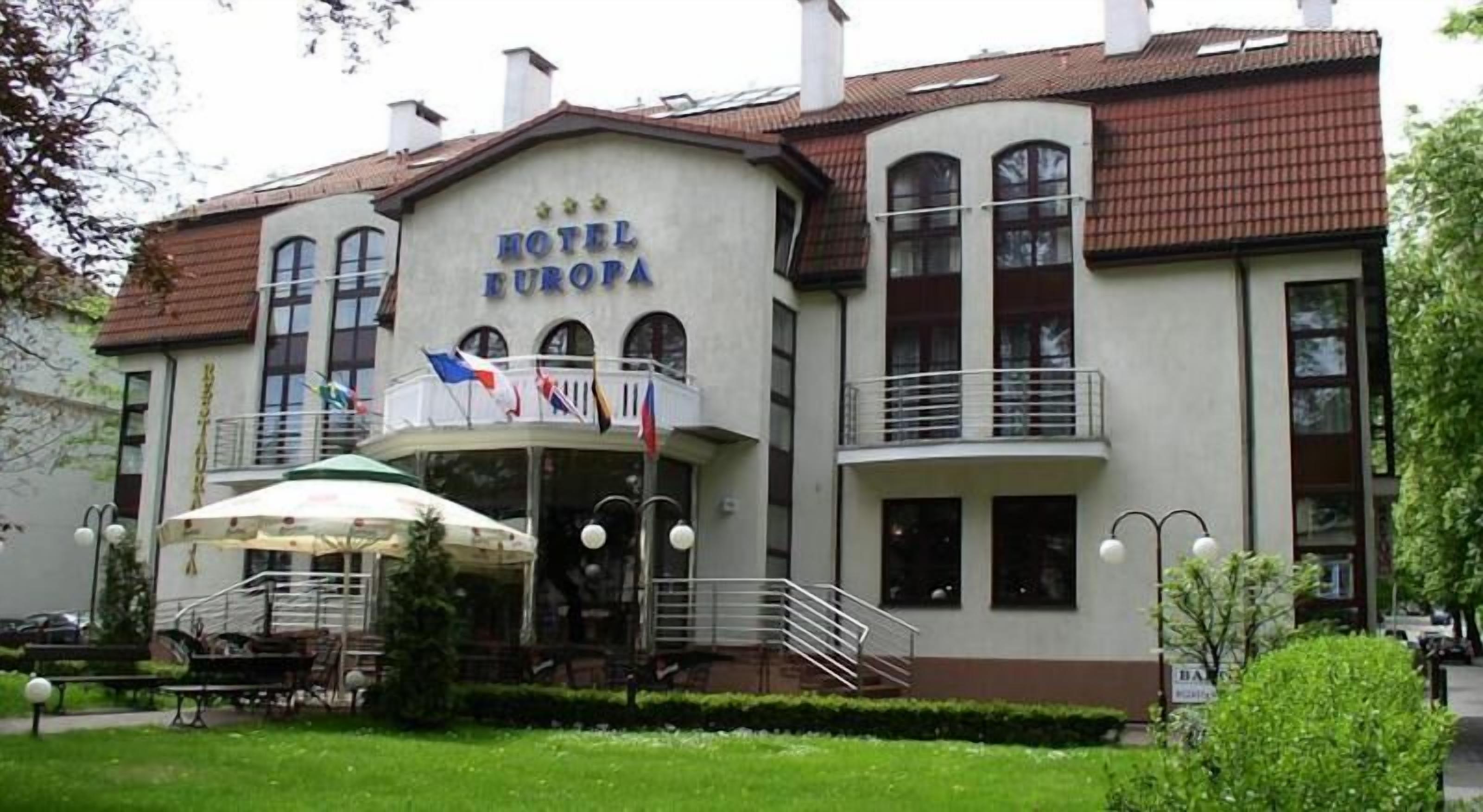 Un hotel con un cartel que dice "HOTEL EUROPA". Tiene un balcón con barandillas, una entrada cubierta con sombrillas y una escalera que conduce a la entrada.