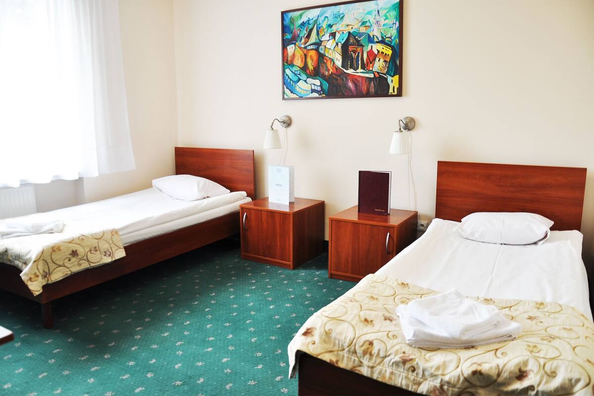A hotel room with two beds, a wooden headboard, a green patterned carpet, a painting on the wall, and a bedside table with a lamp.
