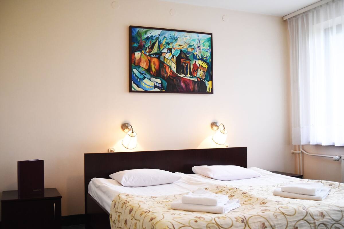A hotel room with a bed, two bedside lamps, a painting on the wall, and a window with curtains.