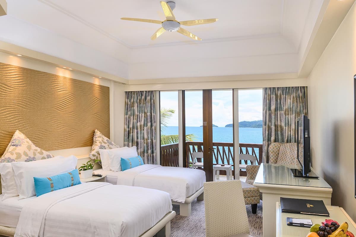 A hotel room with two beds, a desk, a chair, a television, and a large window offering a view of the sea.