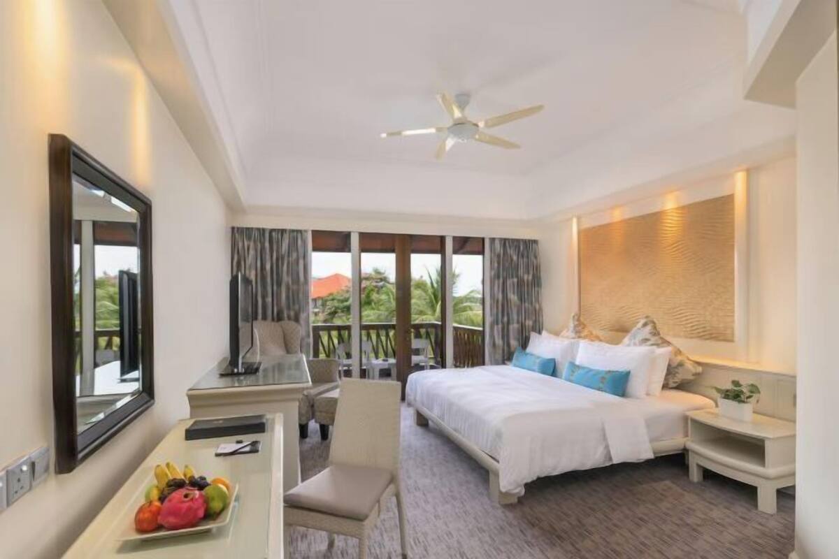 A hotel room with a bed, a desk, a chair, a fruit bowl, and a view of the outdoors.