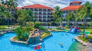 A resort pool area with multiple water features, including a waterfall and slides, surrounded by palm trees and a multi-story building with red roofs.