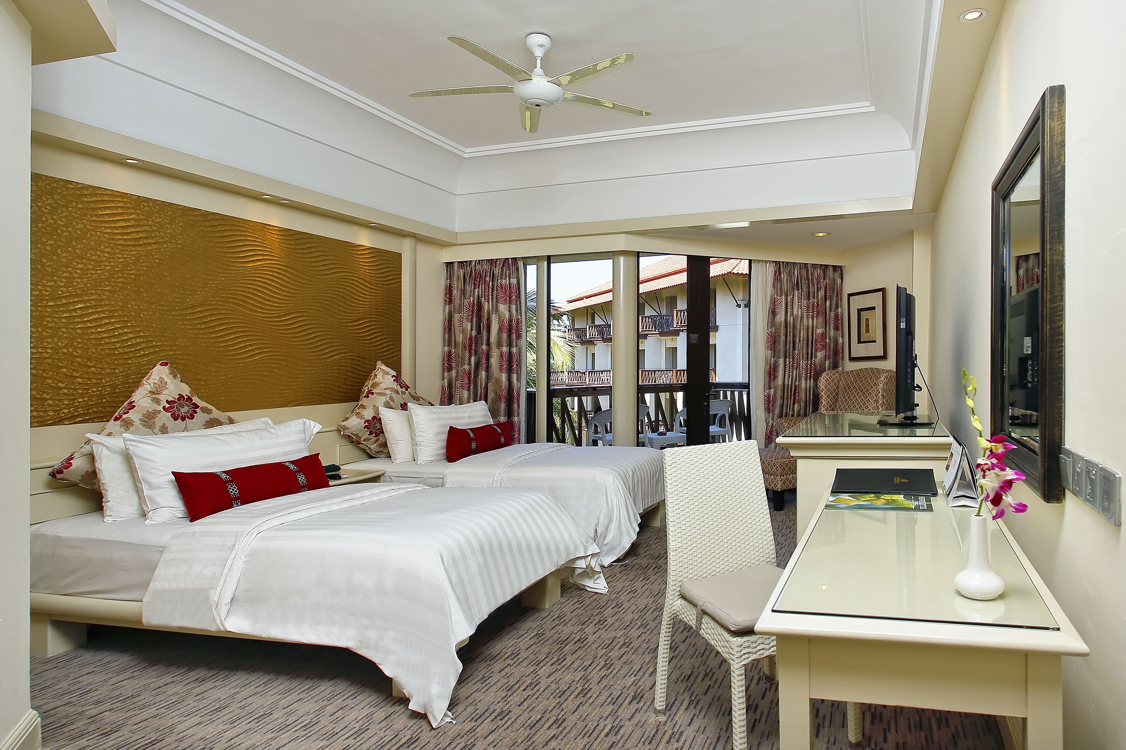 A hotel room with two beds, a desk, a chair, a ceiling fan, and a view of the outside balcony.