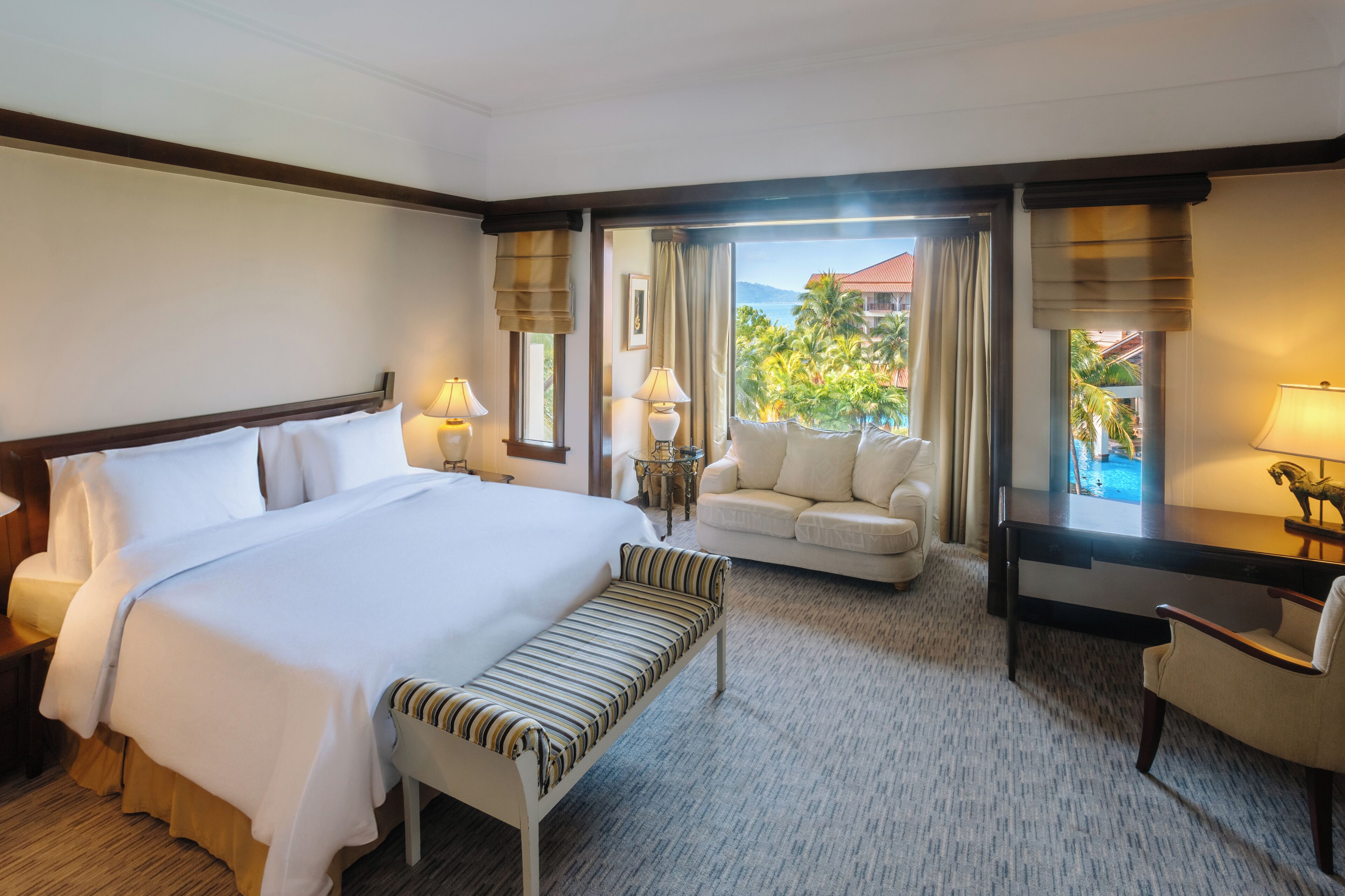 A hotel room with a large bed, a sofa, a desk, and a view of a tropical landscape.