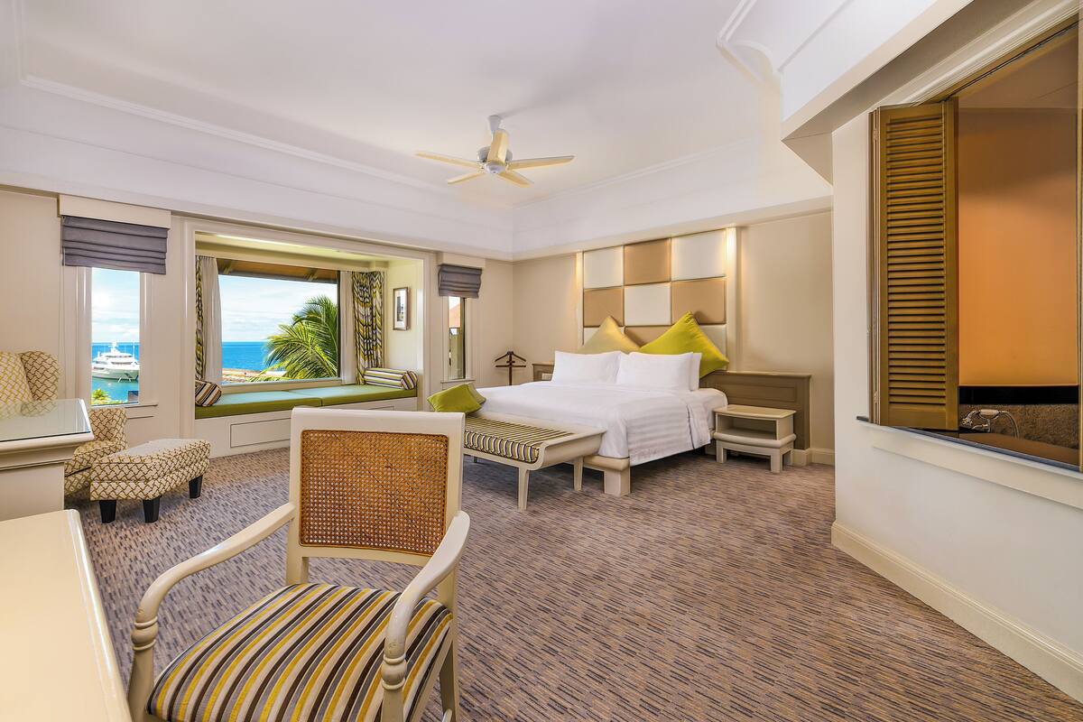 A hotel room with a bed, a desk, a chair, a window with a view of the ocean, and a ceiling fan.