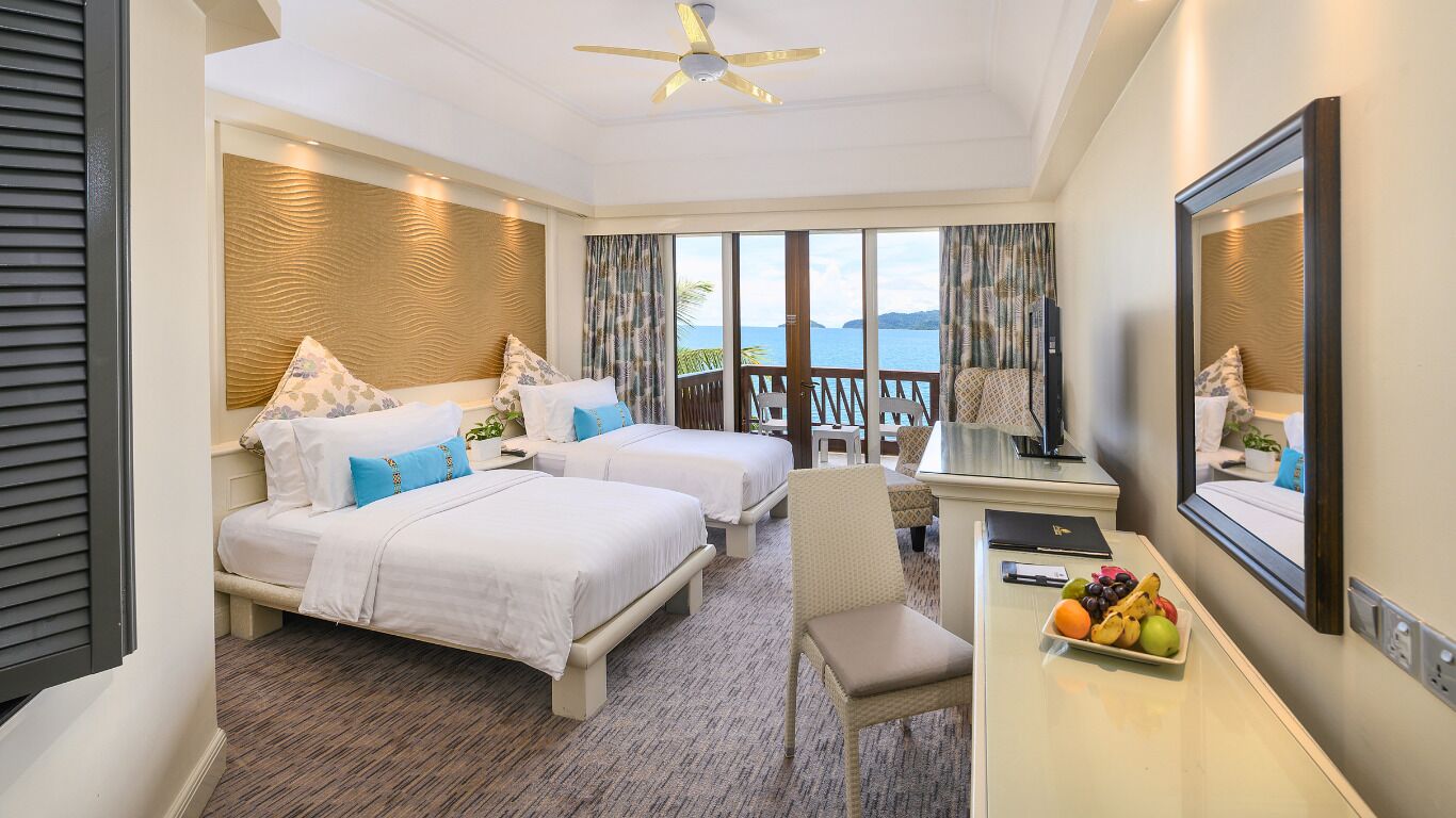 A hotel room with two beds, a desk with a fruit bowl, a chair, and a large window with a view of the sea.
