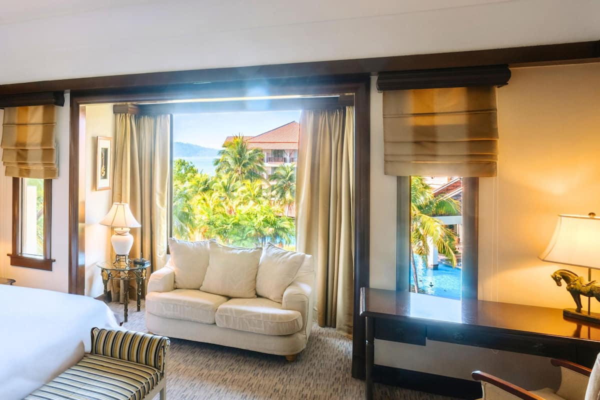 A hotel room with a large bed, a sofa, a desk, and a view of a tropical landscape.