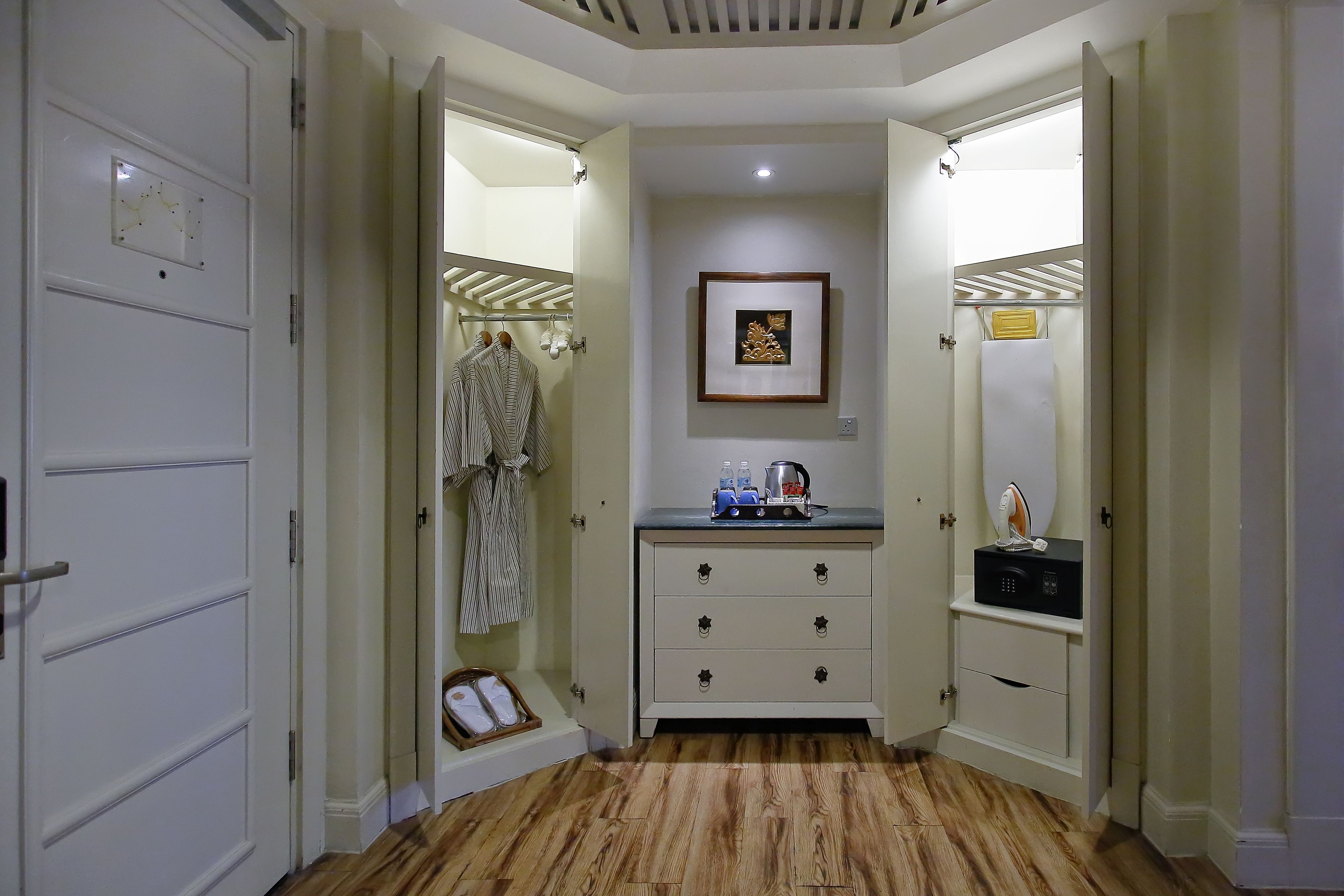 A hallway with a wooden floor, a white door, a coat rack with a striped robe, a framed picture on the wall, and a small table with a kettle and bottles.