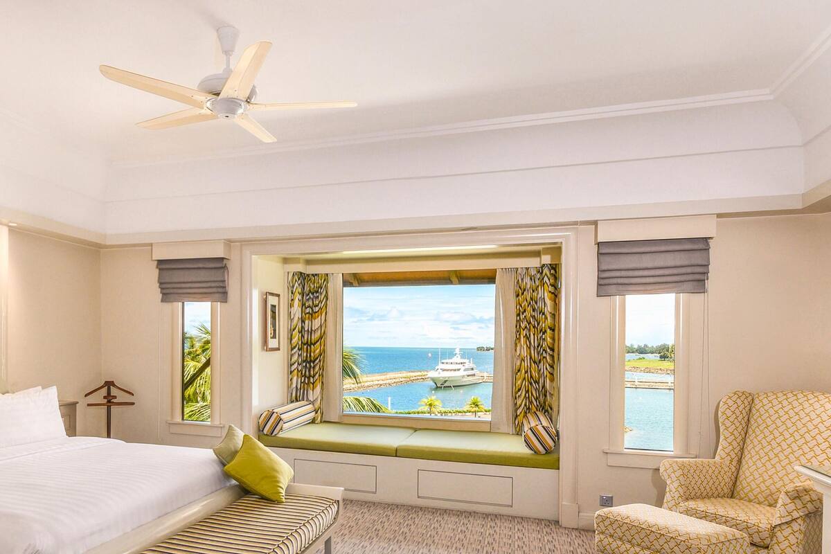 A hotel room with a bed, a seating area with a view of the sea, a ceiling fan, and a window with curtains.