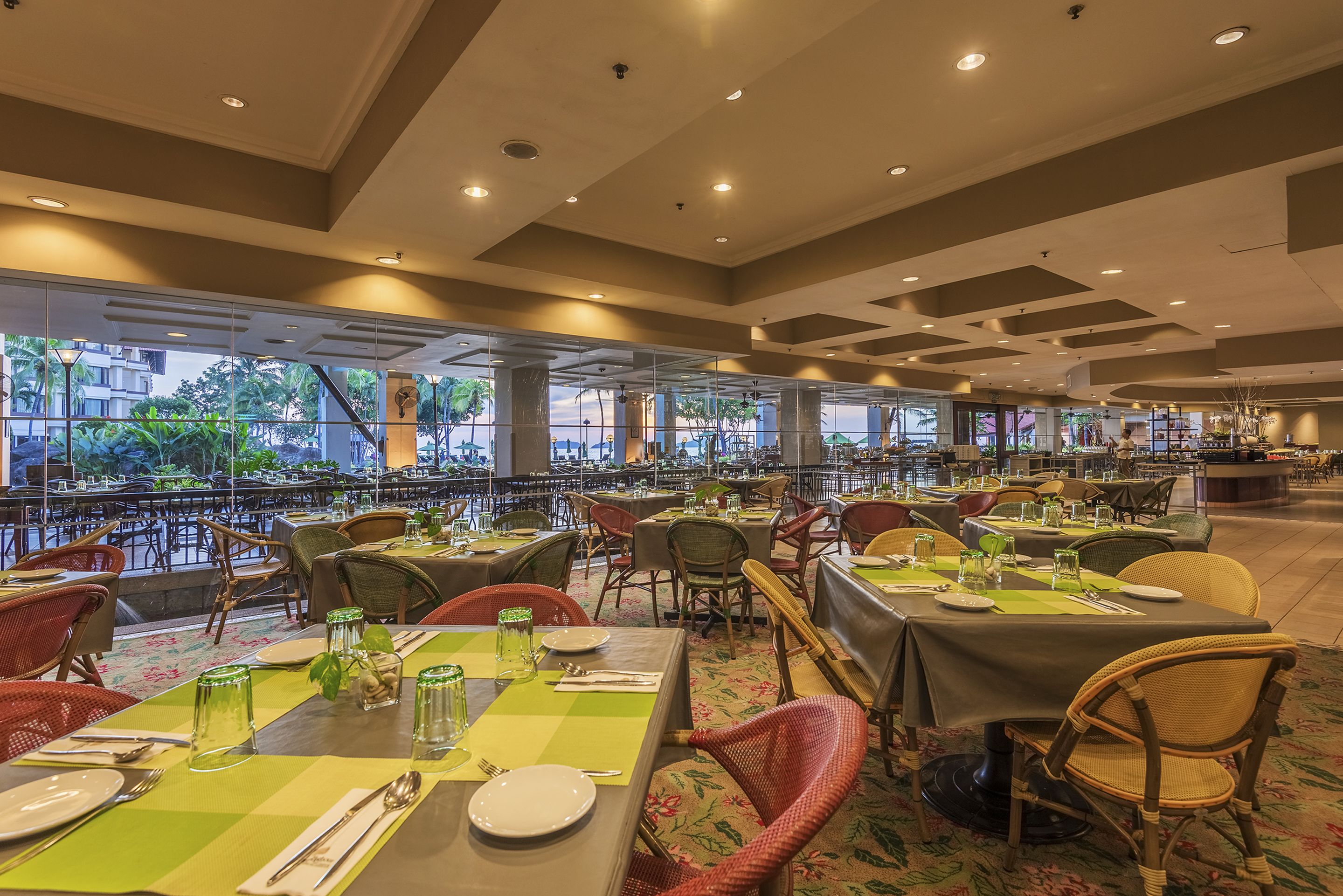 A spacious restaurant with multiple dining tables and chairs, large windows offering a view of the outdoors, and a modern ceiling design.