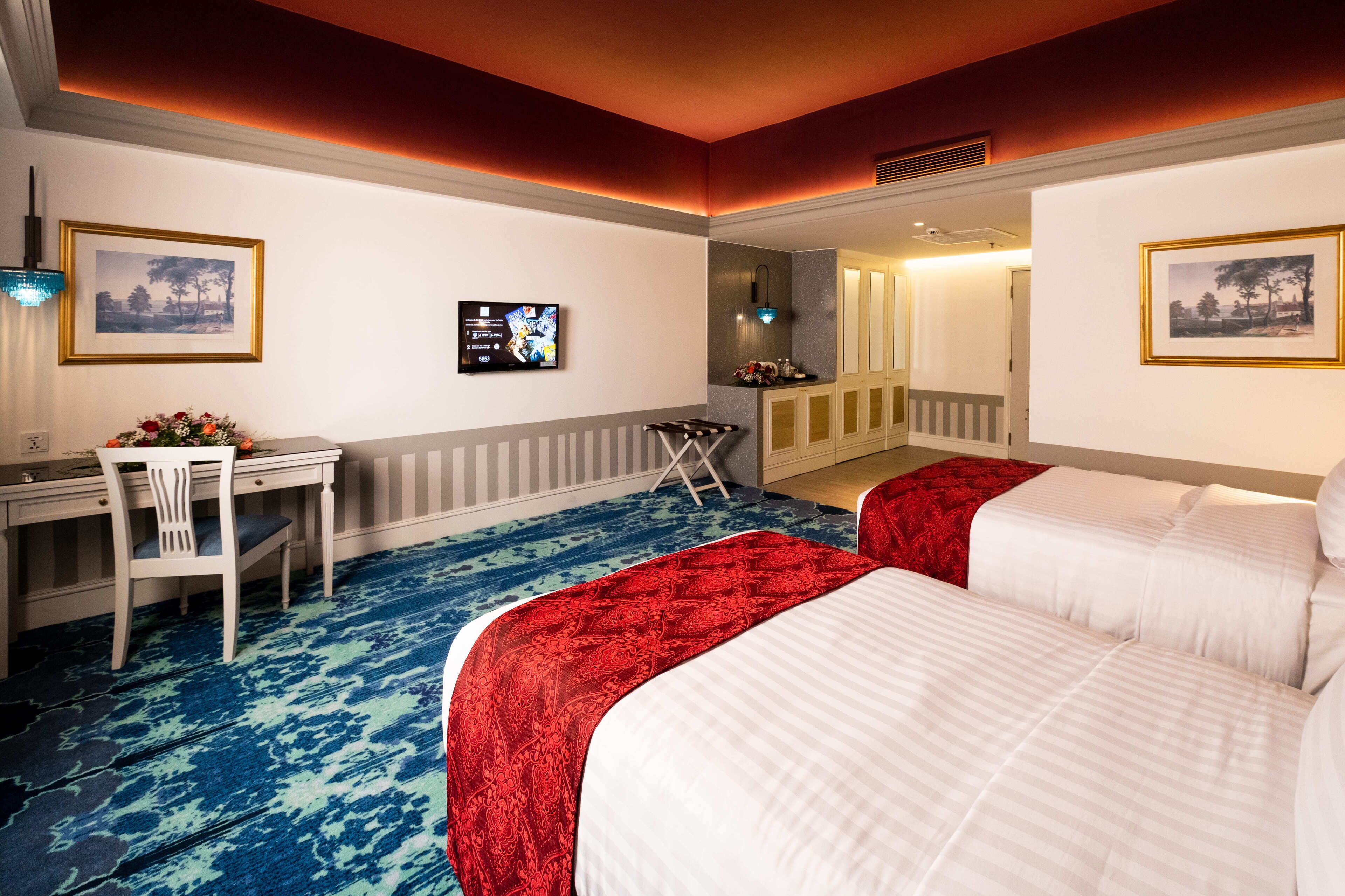 Habitación de hotel con dos camas, un escritorio, una silla, un televisor y obras de arte en las paredes.