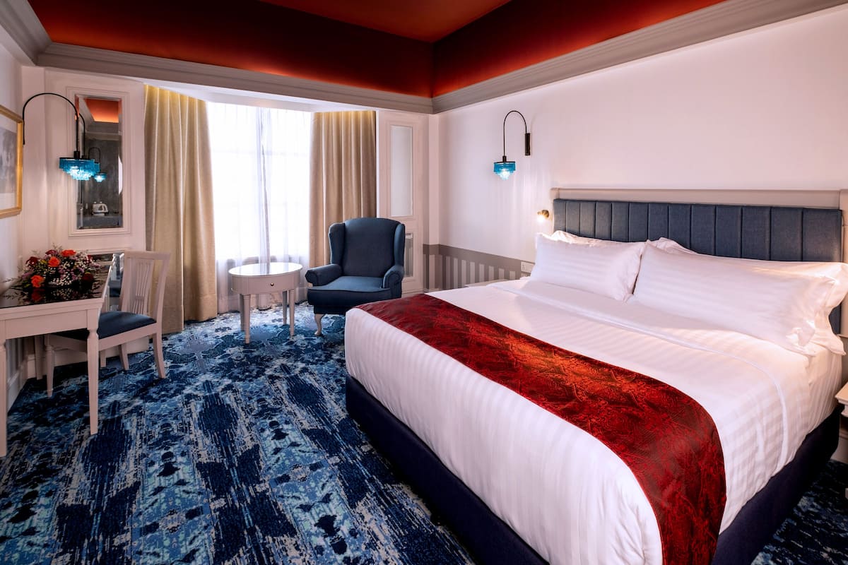 Una habitación de hotel con una cama grande, un escritorio con silla, una mesita pequeña y dos lámparas de pared.