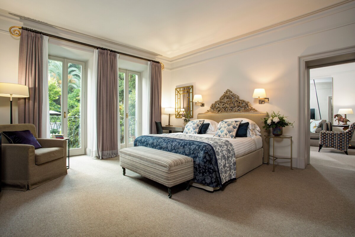 Un dormitorio amplio con una cama grande, una zona de estar con un sillón y vistas a zonas verdes por la ventana.