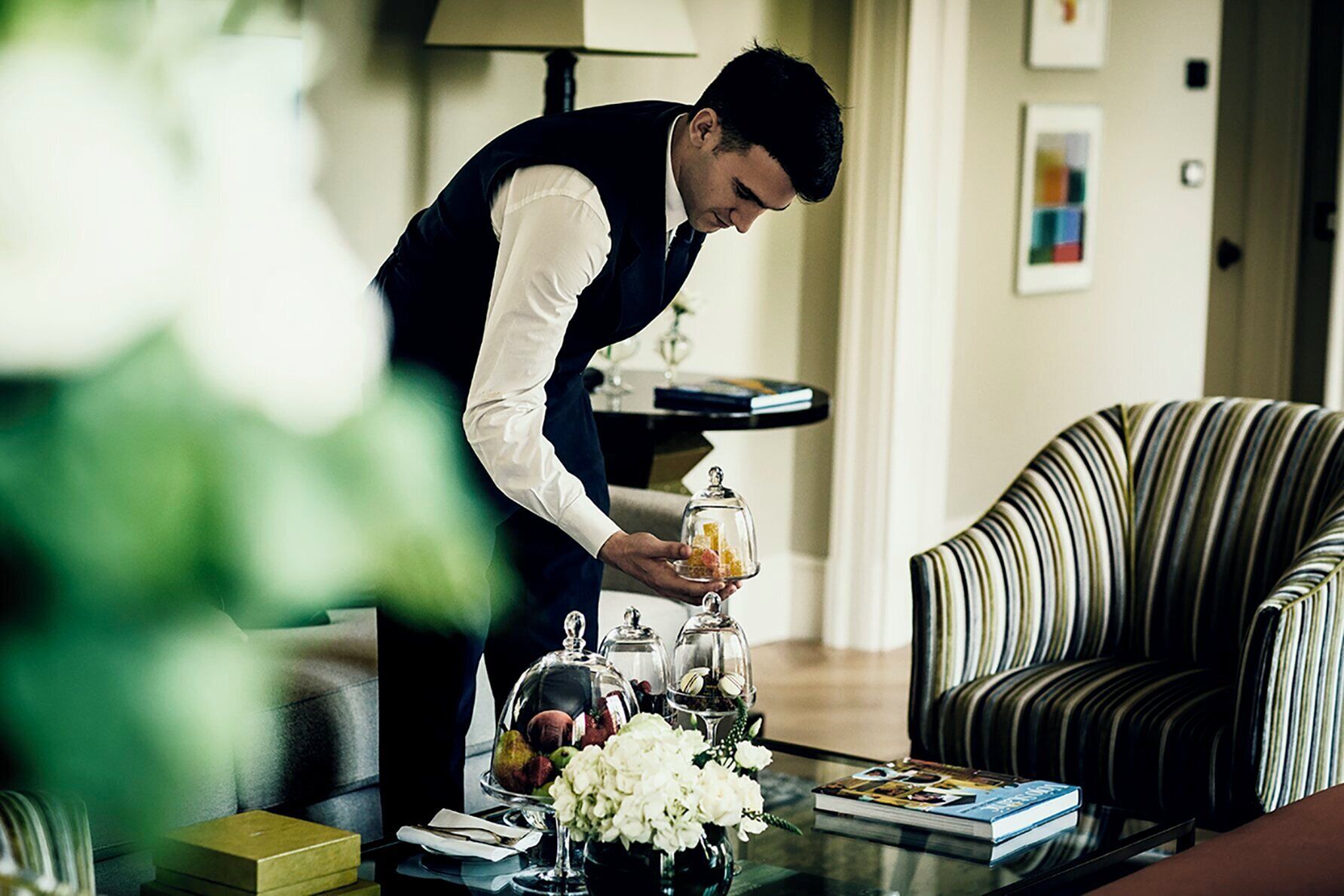 A person arranging a table with a glass dome, a vase of flowers, and a book.