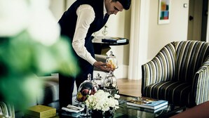 A person arranging a table with a glass dome, a vase of flowers, and a book.