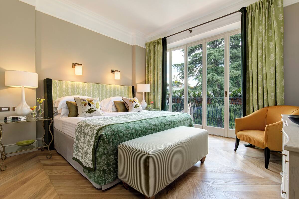 A hotel bedroom with a large bed, two bedside lamps, a bench, a chair, and a view of trees through a sliding door.