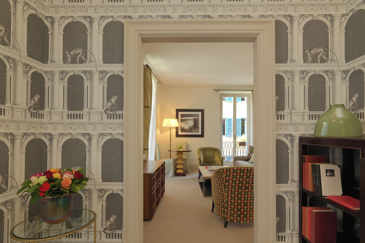 A hallway with a patterned wallpaper, a glass table with flowers, and a view into a room with a chair and a lamp.