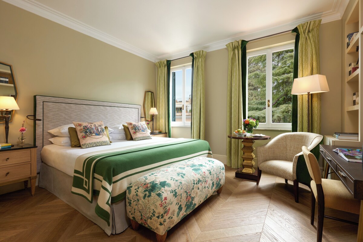Una habitación de hotel con cama, mesitas de noche, escritorio, silla y ventana con cortinas.
