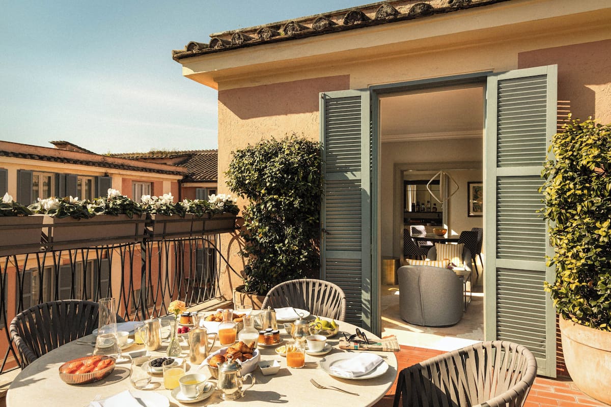 A round table set for breakfast with various dishes and drinks, surrounded by chairs, in an outdoor patio area with a view of a building and a potted plant.