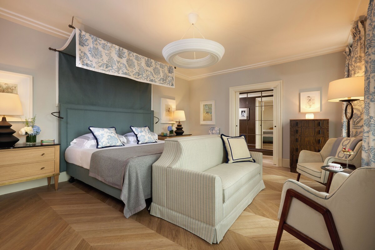 Un dormitorio amplio con una cama grande, una zona de estar con sofá y sillones, un aparador de madera y un ventanal.