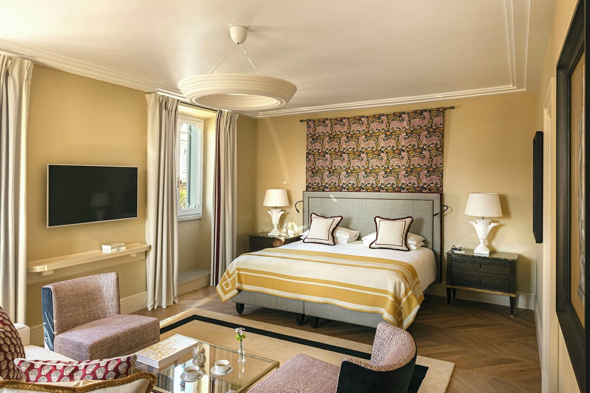 Habitación de hotel con cama, dos mesitas de noche, televisor, ventana con cortinas, un tapiz estampado y una zona de estar con mesa y sillas.