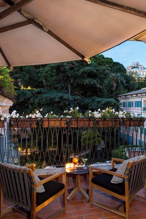 An outdoor patio with two wooden chairs, a table with candles, and a view of a garden and buildings in the background.