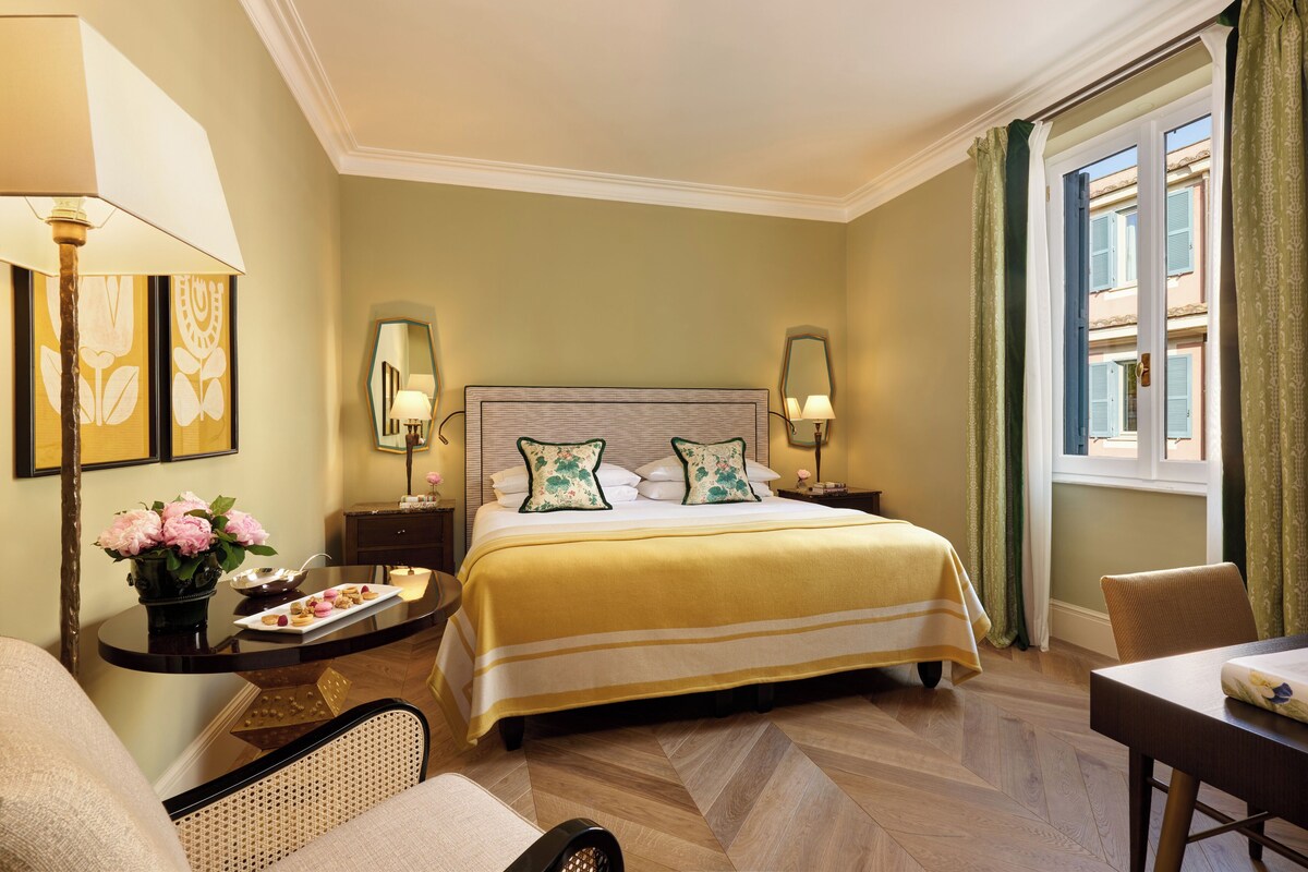 A hotel room with a large bed, two bedside tables with lamps, a small table with a vase of flowers, a chair, and a view of the outside through a window.