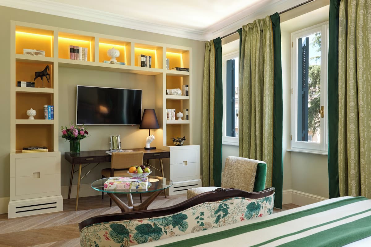 A hotel room with a large flat-screen TV, a desk with a lamp, a chair, a sofa, and a bookshelf with various items.