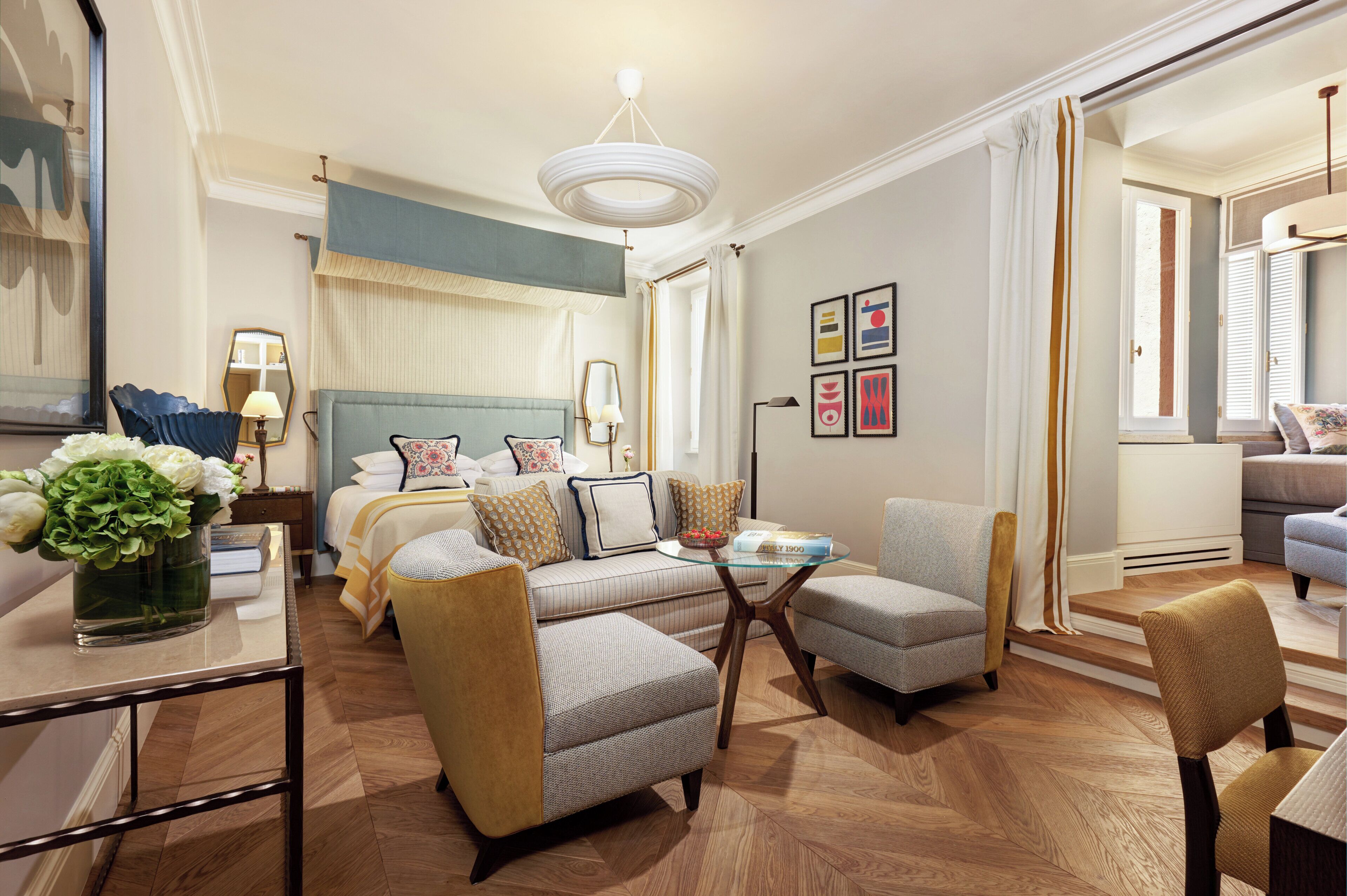 Quarto de hotel com uma cama, área de estar com uma mesa, estante de livros e uma grande janela com cortinas.