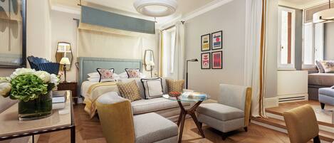 Quarto de hotel com uma cama, área de estar com uma mesa, estante de livros e uma grande janela com cortinas.