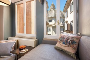 A cozy bedroom with a large window, a bedside table with a fruit bowl, and a decorative pillow.