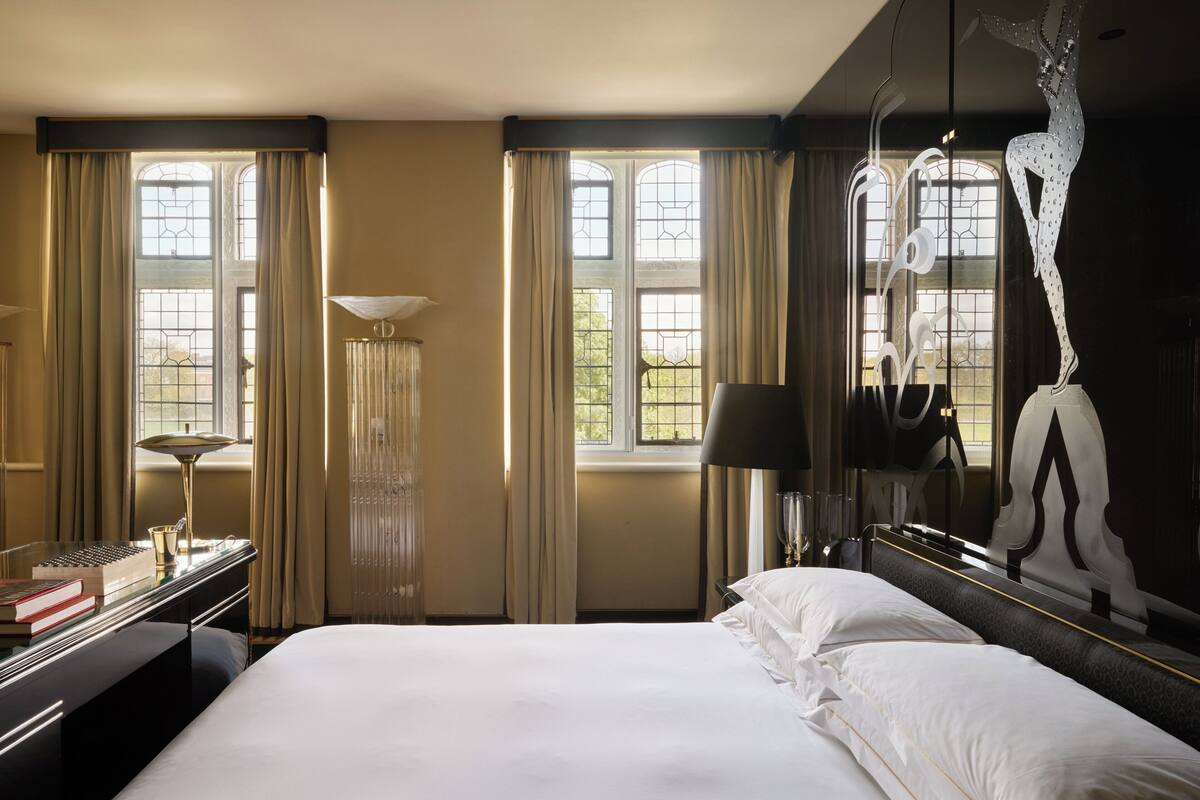 A hotel room with a large bed, a desk with a lamp, and a view of the outdoors through large windows.
