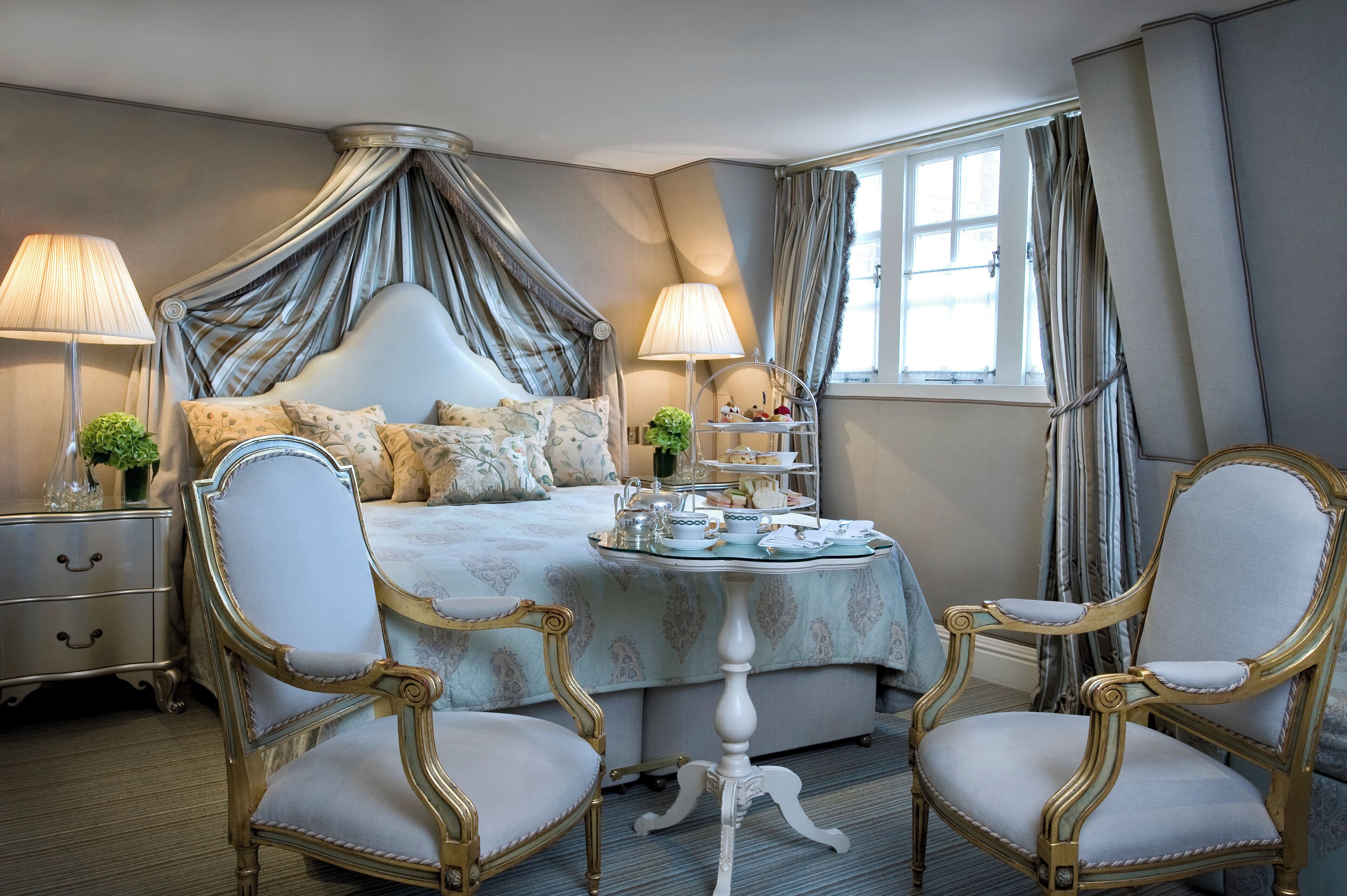 A bedroom with a four-poster bed, two chairs, a small table with tea service, and a nightstand with lamps.