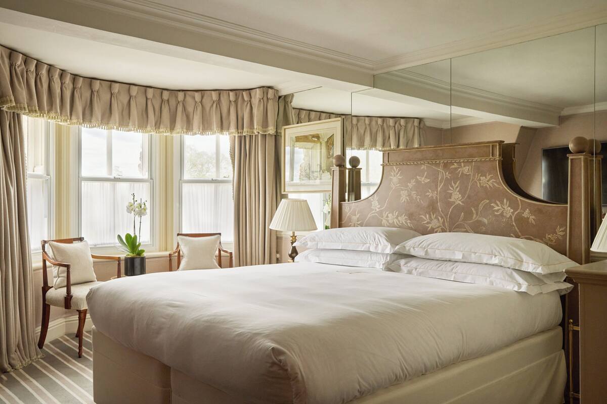 A large bed with white linens, a wooden headboard with floral patterns, a bedside table with a lamp, and a chair with a potted plant.