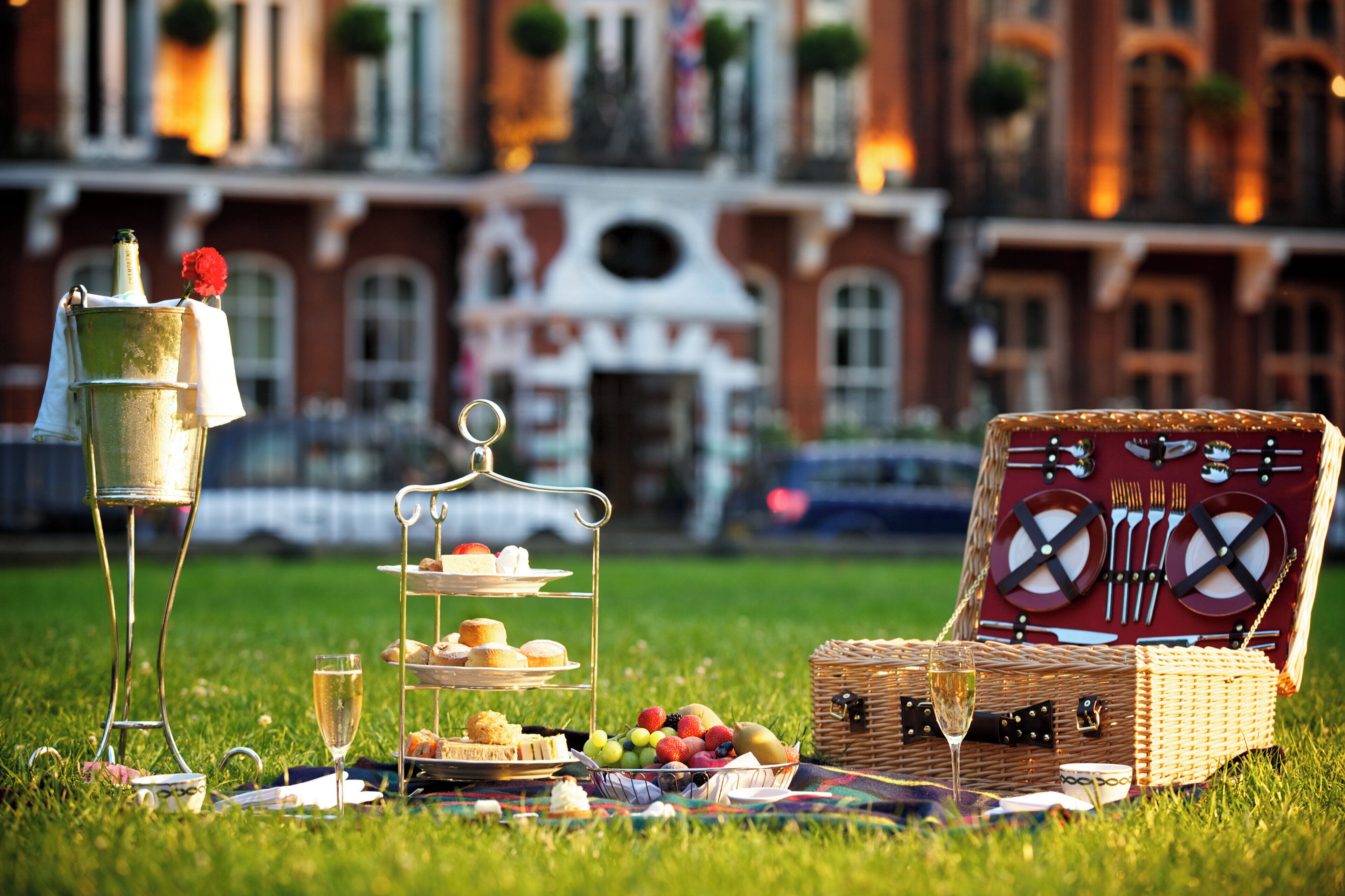 A picnic setup with a three-tiered stand, a wicker picnic basket, and a metal stand with a bottle and a rose.