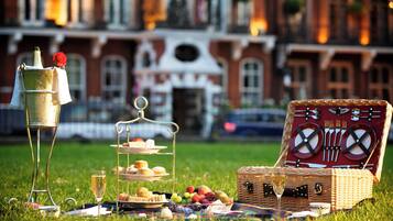 A picnic setup with a three-tiered stand, a wicker picnic basket, and a metal stand with a bottle and a rose.