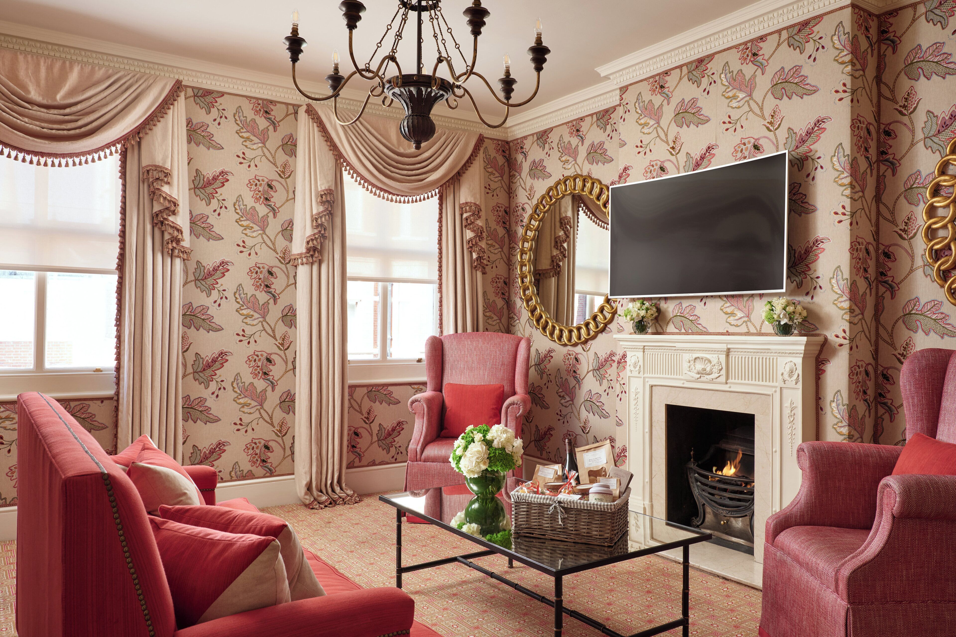 Uma sala de estar com lareira, papel de parede floral e mobiliário estofado em vermelho.