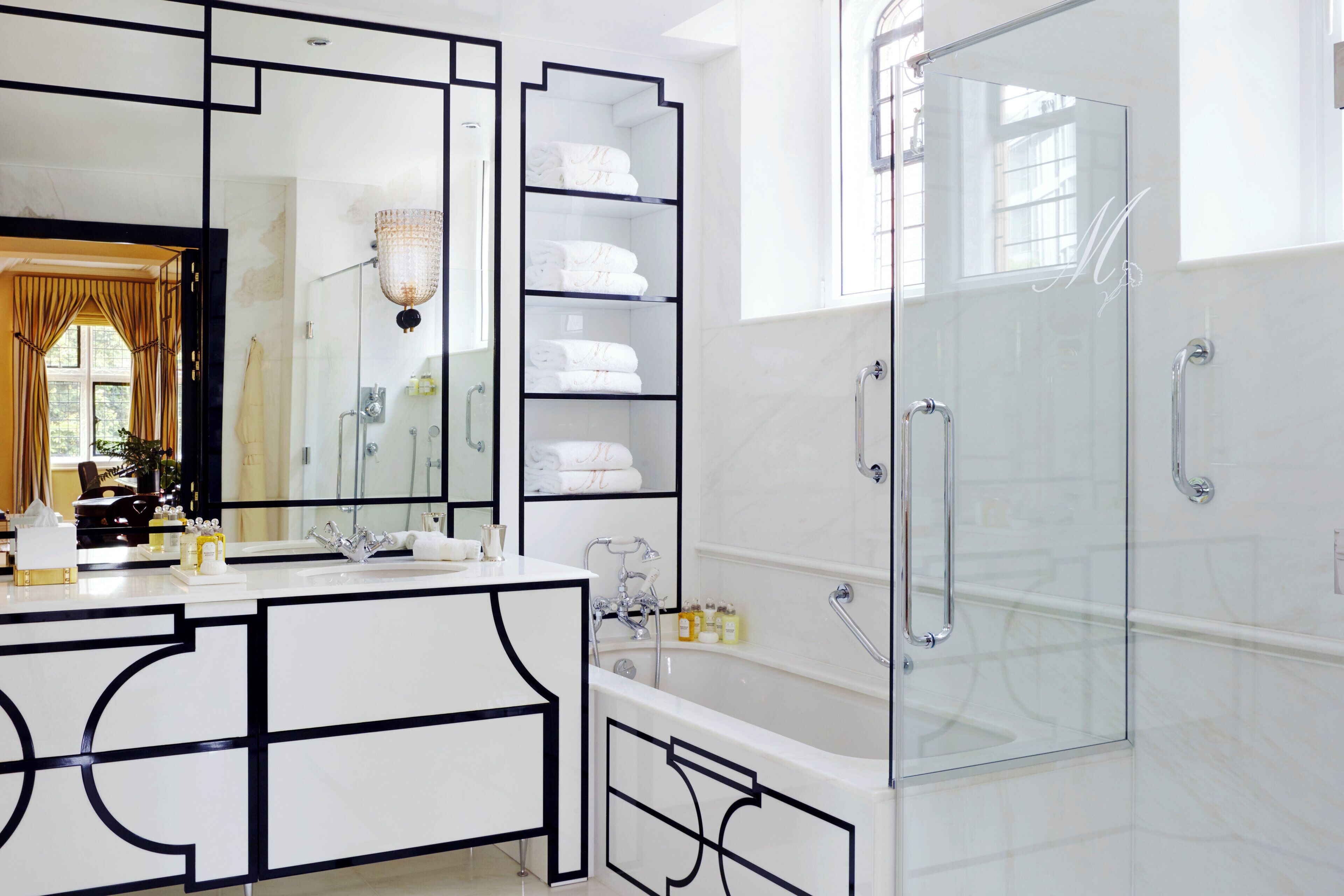 A modern bathroom with a large mirror, white vanity, glass shower enclosure, and built-in shelves with towels.