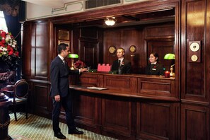 A reception area with a wooden counter, a desk, and a chair. There are clocks on the wall and a vase with flowers on the left. - The Milestone Hotel (London)