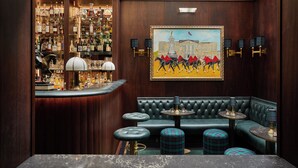 Zona de bar con mostrador de madera, estanterías llenas de botellas, un cuadro de una carrera de caballos y un sofá de cuero verde con otomanas a juego.