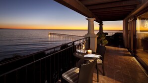 A balcony with a view of the ocean at sunset, featuring a table with a candle, a chair, and a potted plant.
